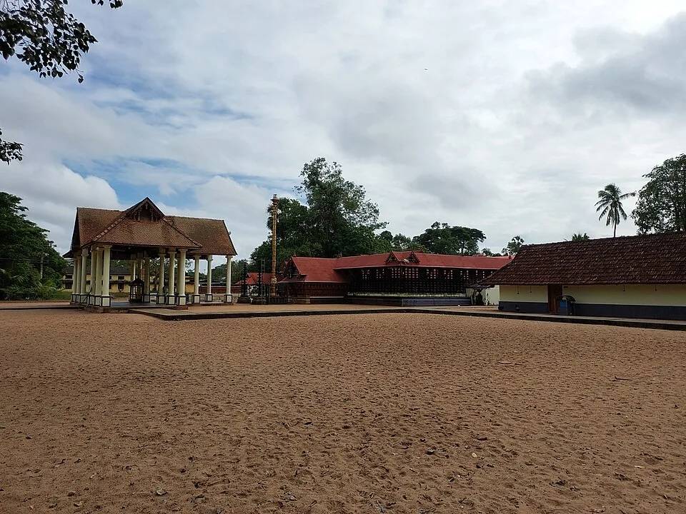 A very wide expanse of red laterite sand inside a temple compound, with a pillared mandapam on the left, a tall flagstaff and the tiled temple buildings beyond.