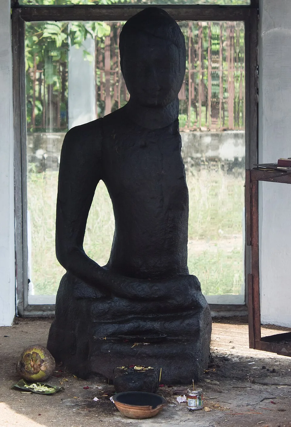 The Karumadikuttan Buddha with its left arm missing, and on the floor before it a split coconut on a banana leaf, loose flowers, a brass bowl and a stone lamp