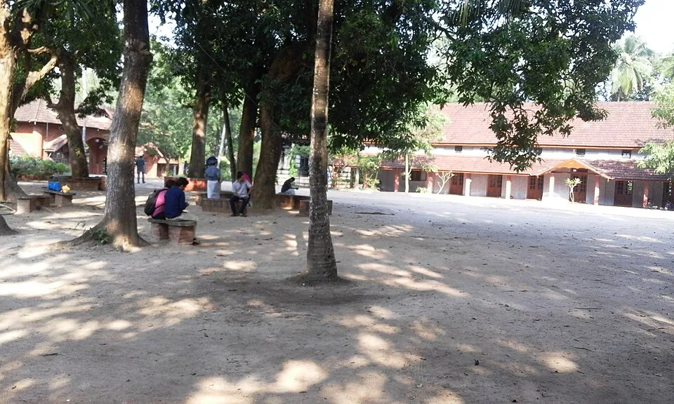 Open sandy ground under large trees inside the Thunchan Memorial grounds at Tirur