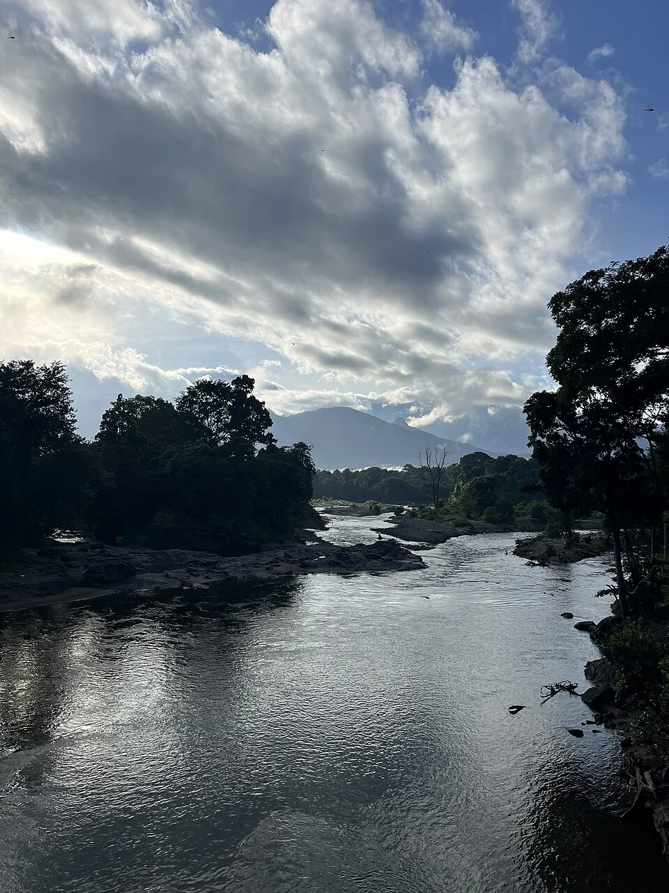 The Karimpuzha flows between wooded banks and exposed rocks below cloud and distant hills at Nedumkayam.