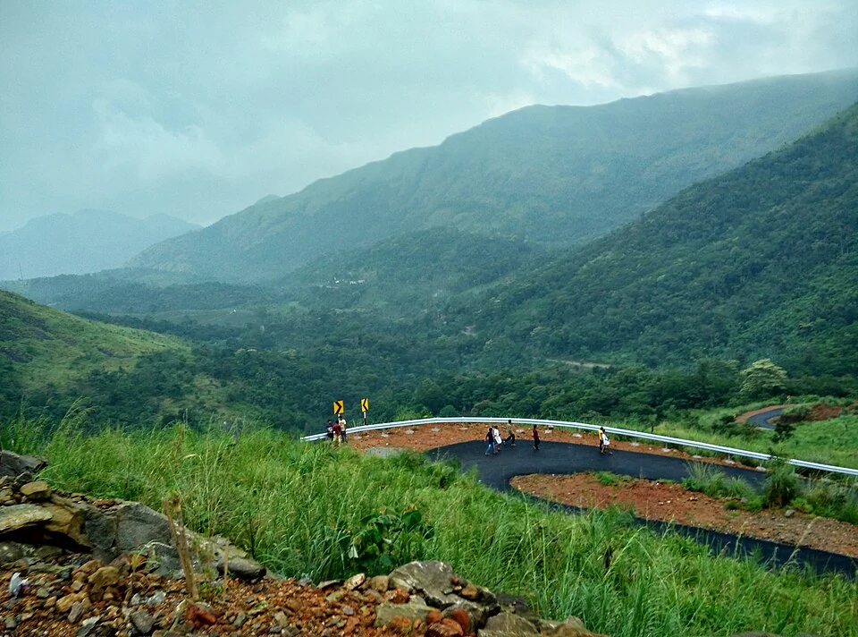 Visitors walk on a tight paved bend with a metal guardrail below the green slopes of Illikkal Kallu.