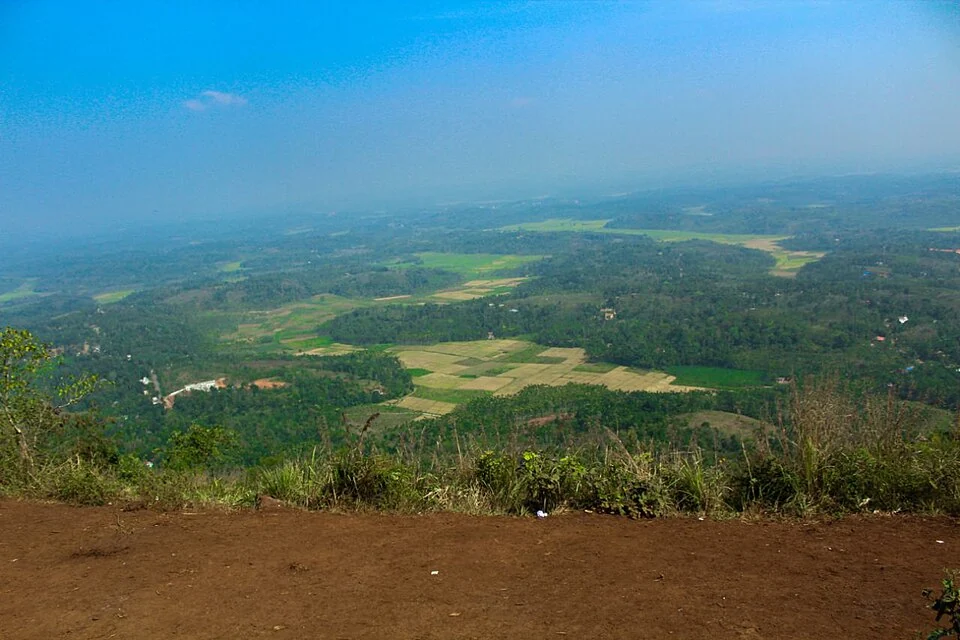 Patchwork fields and tree-covered land stretch below the earth-edged viewpoint at Kurumbalakotta.