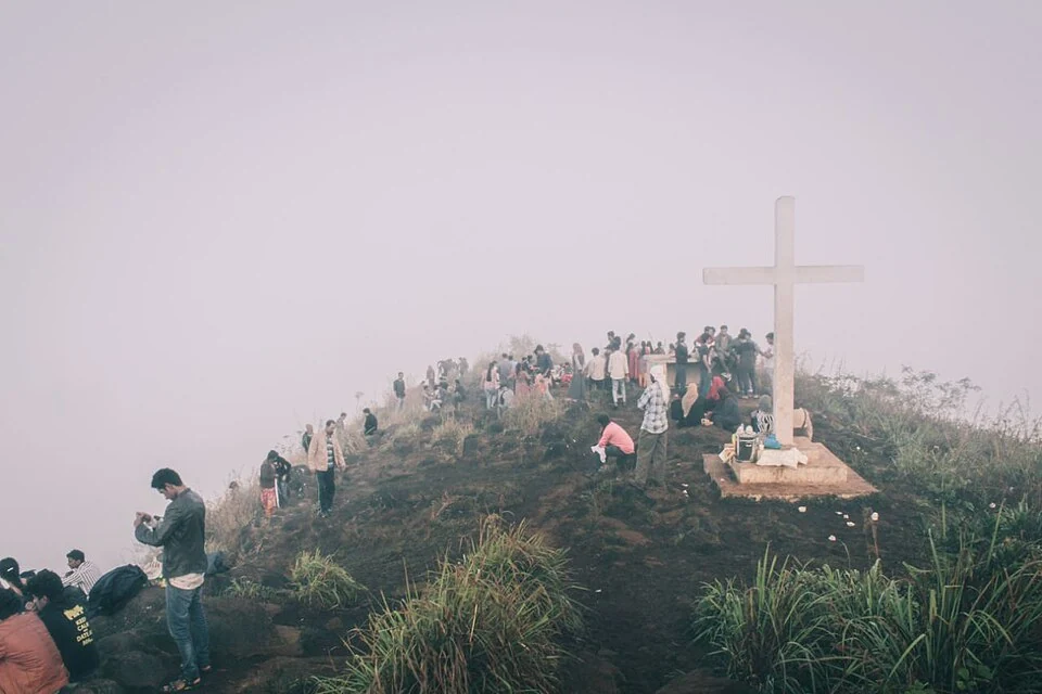 Visitors stand along a mist-covered Kurumbalakotta ridge beside a large white cross.