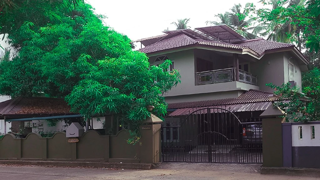 Heavens Homestay stands behind a metal gate, with an upper balcony, tiled roofs and a large tree beside the boundary wall.