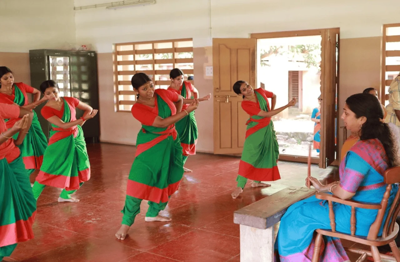 Dance students in green and red practise together while a seated teacher watches from the right.