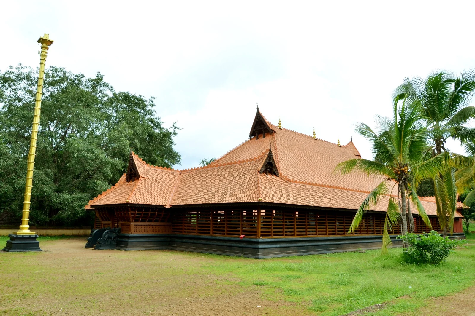 Kerala Kalamandalam’s Koothambalam has layered tiled roofs, timber sides and a dark raised base, with a tall golden flagstaff beside it.
