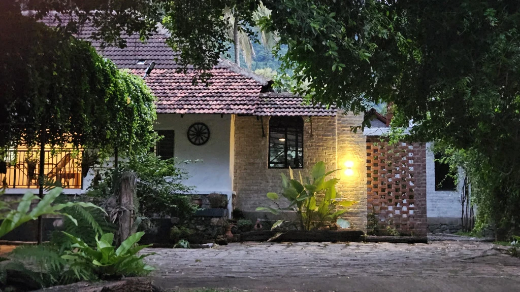 Pannadikadu Homestay’s tiled-roof building stands beneath leafy trees, with warm lights illuminating a stone-faced entrance wall.
