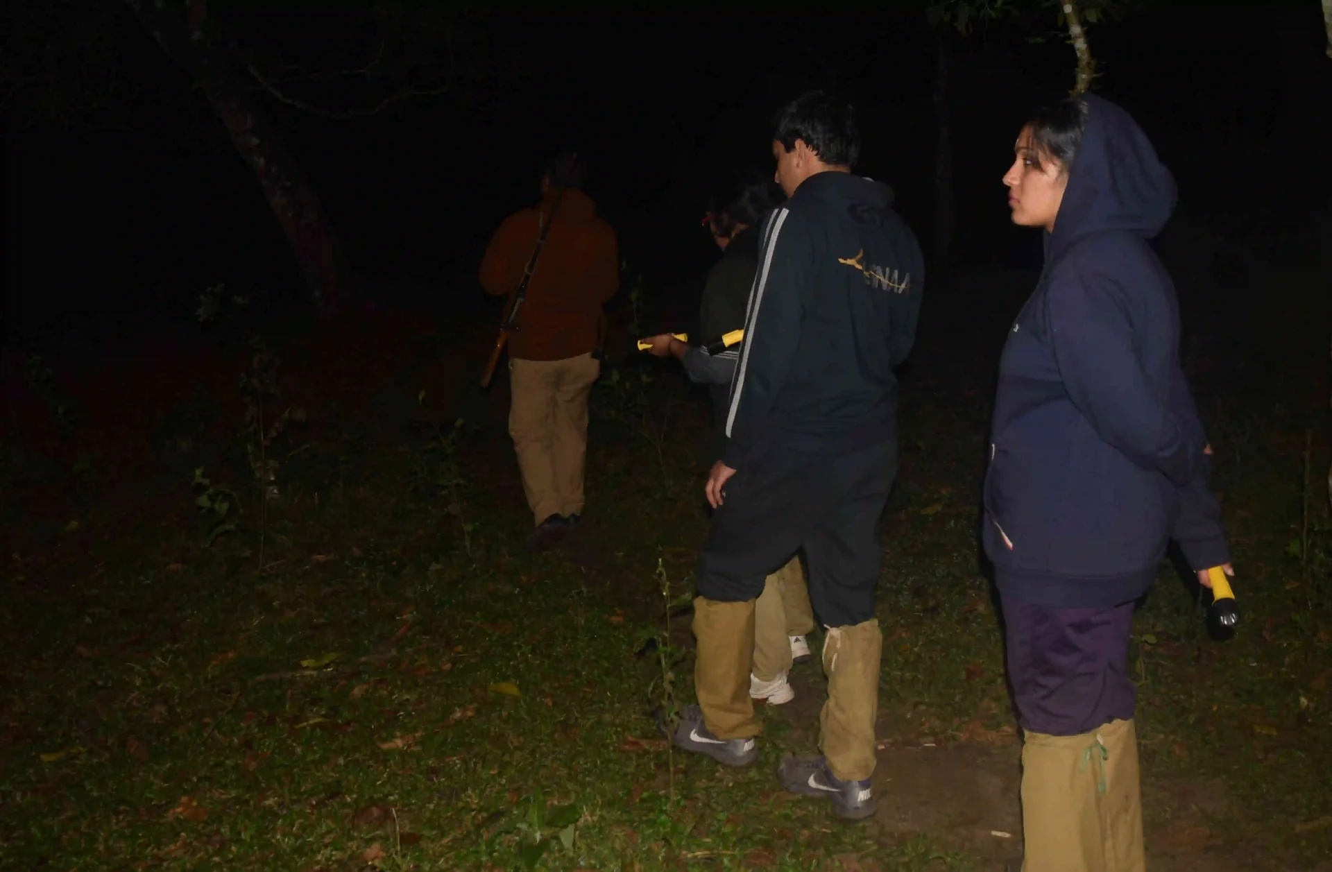 A group carrying torches follows an armed guide along a leaf-covered forest path at night.