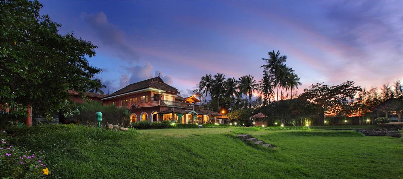 A lit tiled-roof building overlooks the lawn and palms at River Retreat at dusk.