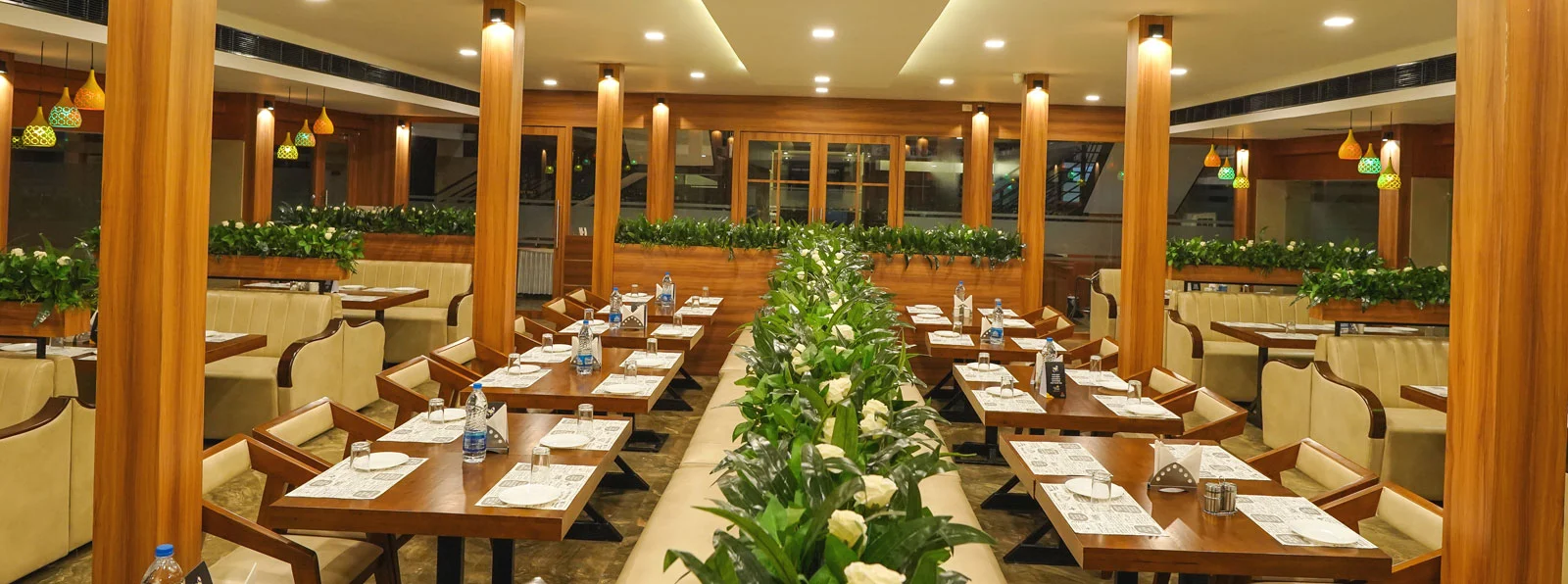 Wooden dining tables and cream upholstered seats line the restaurant, with plants separating the seating areas at Sunstar Residency.