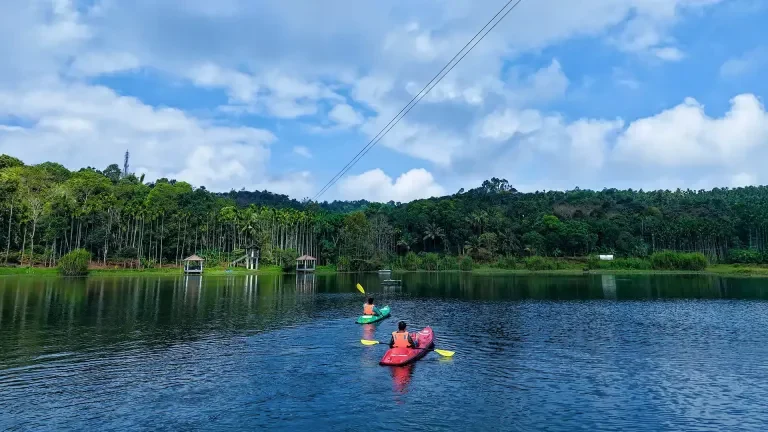 Two kayakers paddle across Karlad Lake beneath overhead cables, with wooded shores beyond.