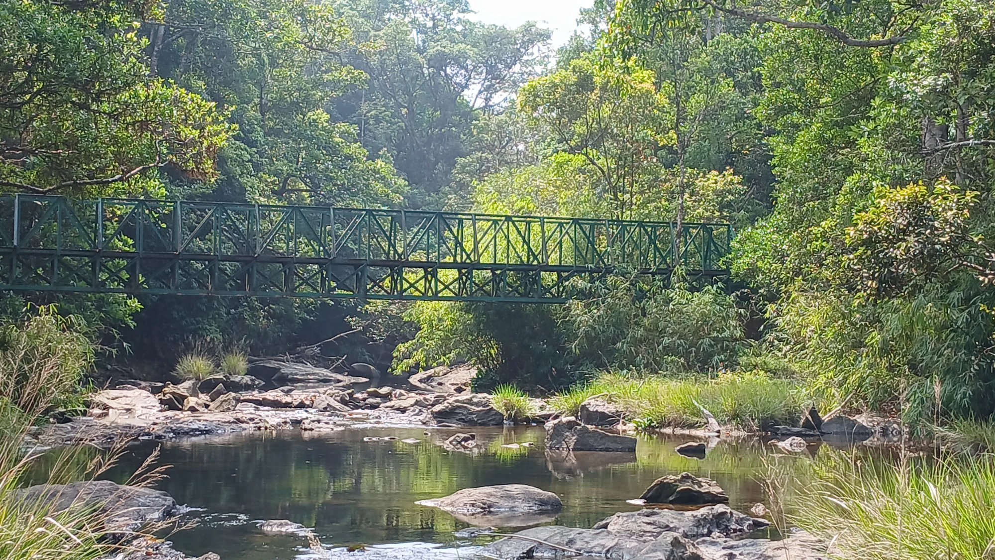 A green metal footbridge crosses a rocky forest stream in the Kakkayam area