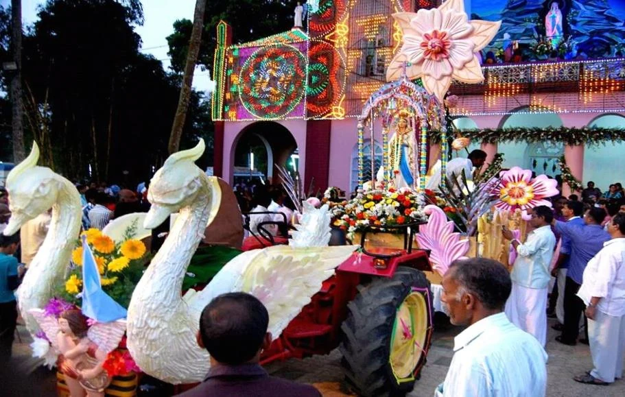 A decorated vehicle carries Marian imagery, flowers and two large white swan figures past Pallikkunnu’s pink church façade.