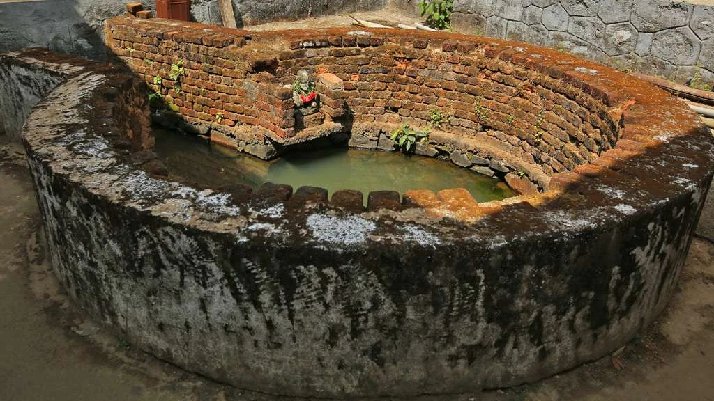A small garlanded figure stands within a low oval water enclosure with weathered brick and stone walls at Thrissilery.