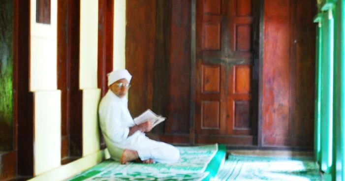 A man in white sits reading beside dark panelled timber doors inside Varambetta’s maqaam.