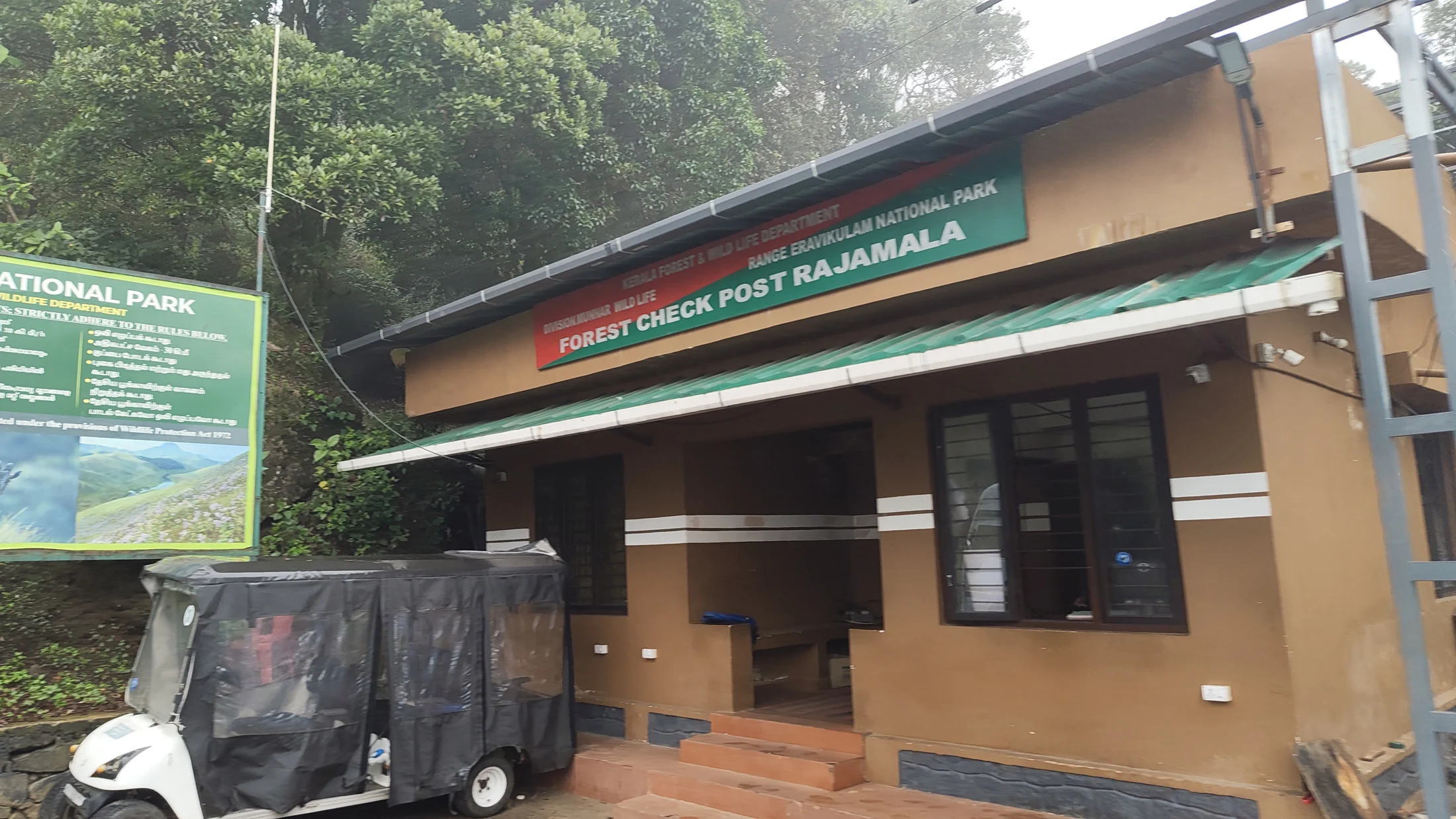 The Forest Check Post Rajamala building has a green sign, a tiled approach and a small electric cart beside it.