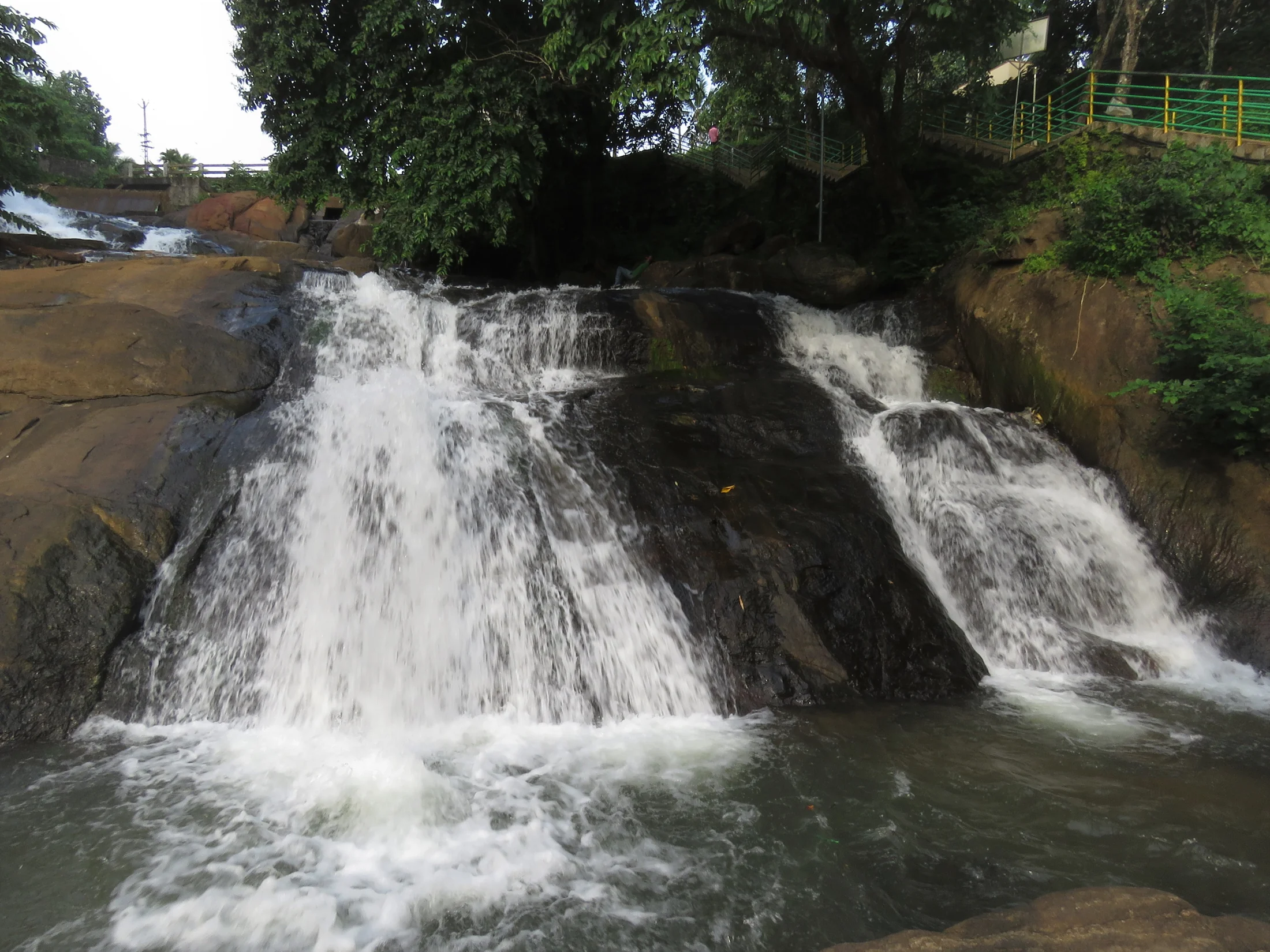 A bridge spans the channel above Aruvikkuzhi’s stepped cascade, with vegetation on both banks.
