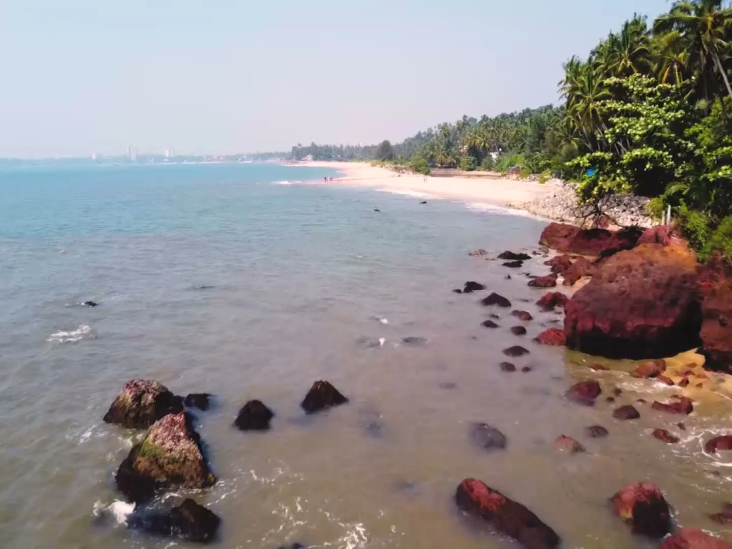 Dark foreground rocks and red coastal outcrops frame Adikadalayi’s sandy shore, with palms extending into the distance.