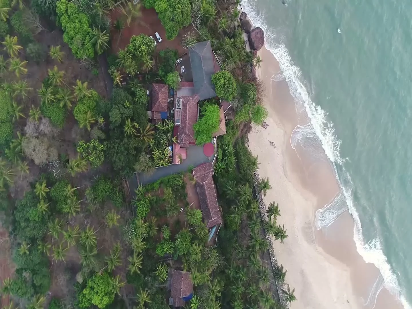 An overhead view shows Seashell Haris Beach Home’s roofs and palms directly above the Adikadalayi shoreline.