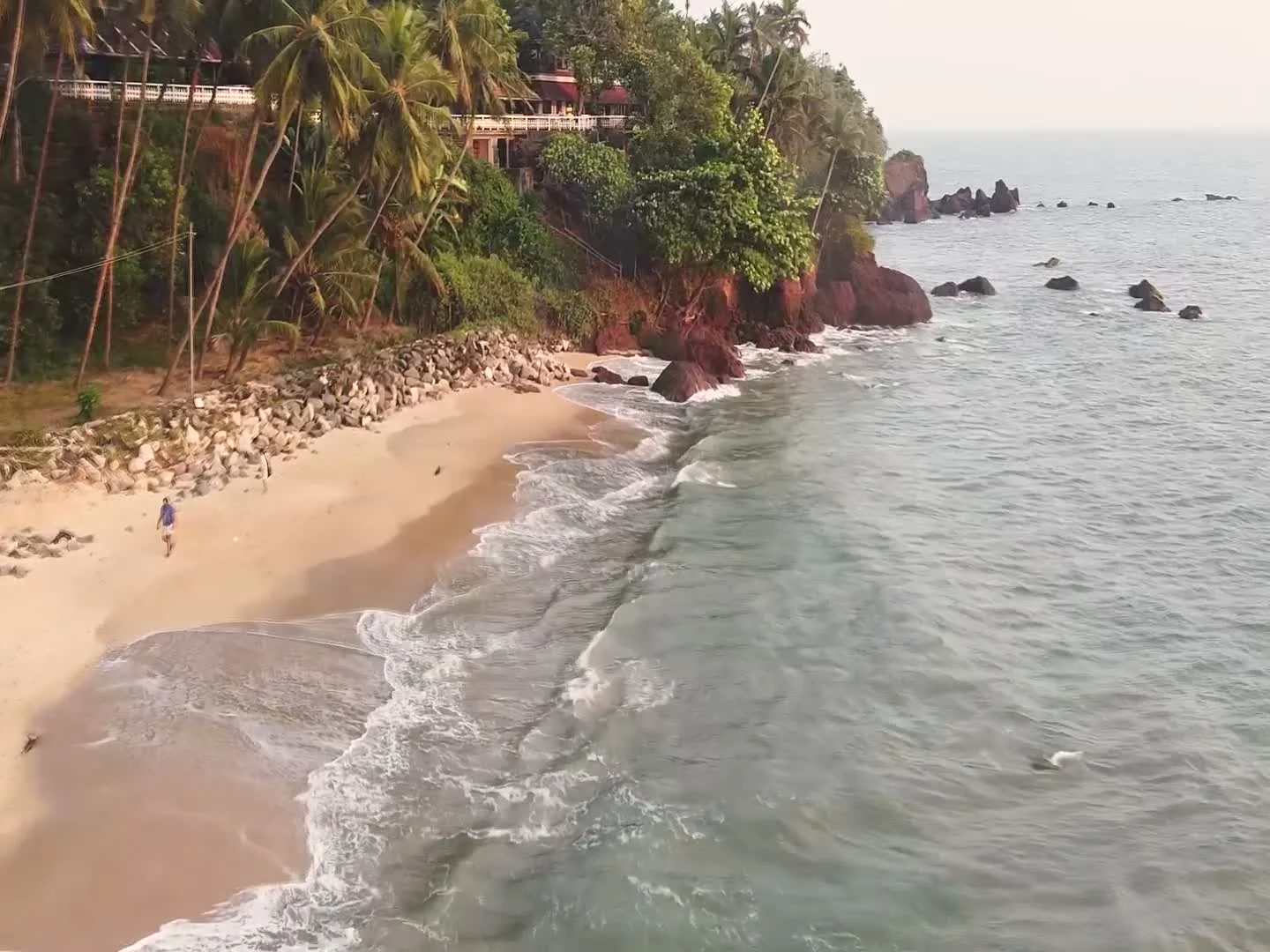 A palm-covered red cliff and stone revetment rise above Adikadalayi’s narrow shore, with a homestay terrace above.