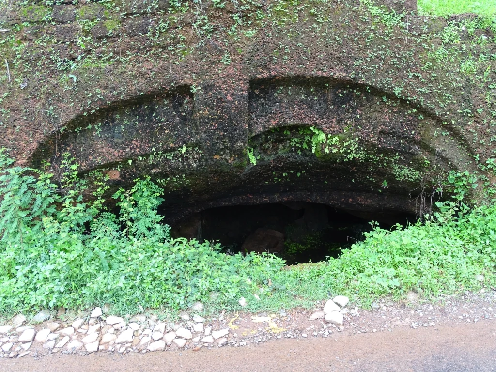 A low opening cuts into a fern-covered laterite bank in Kerala Tourism’s Kanhangad cave-fort photograph.