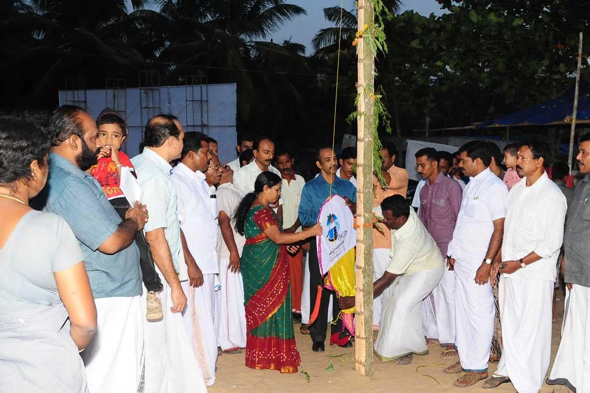 People gather around a decorated flagpole on Nattika’s sand at dusk in an archival festival photograph.