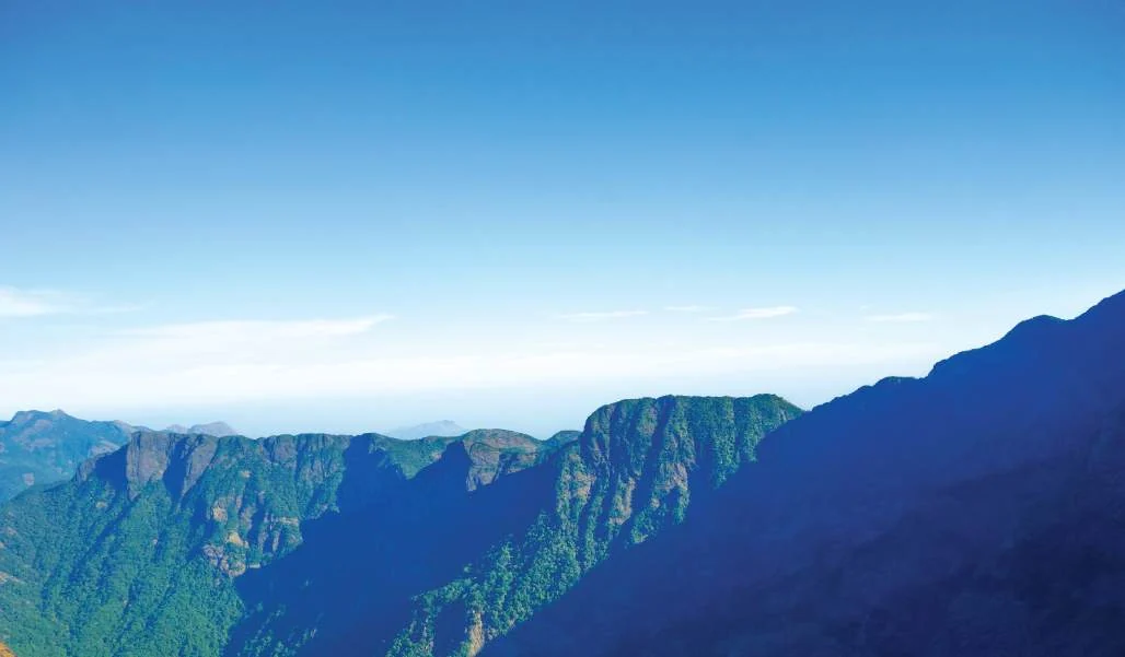 Sunlit cliffs and forested slopes rise beneath a blue sky in the Vellarimala range.